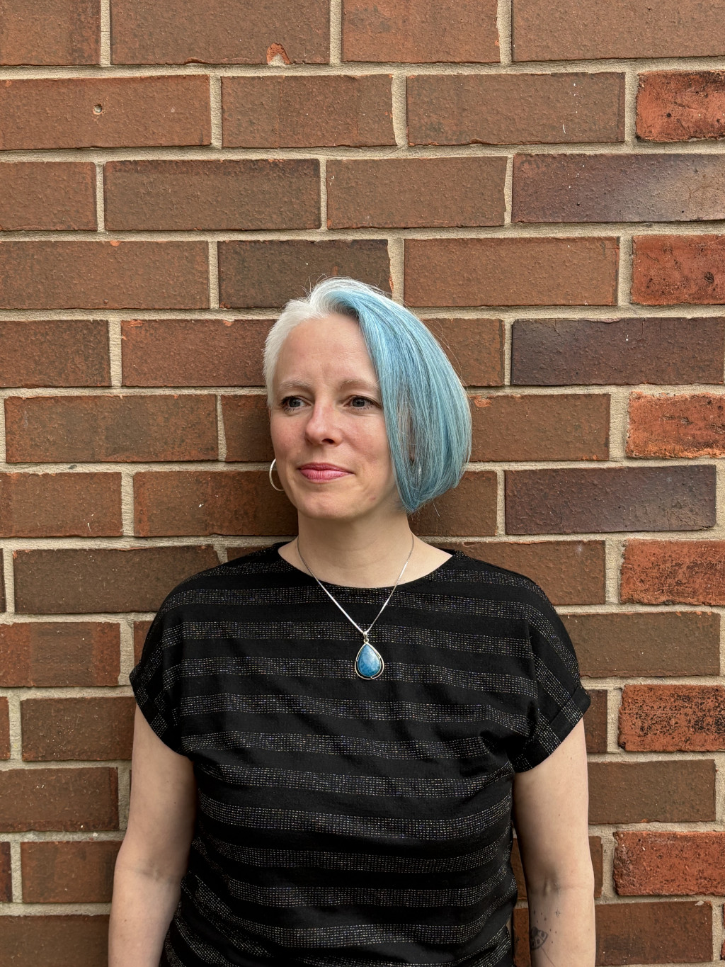 Diny with asymmetric blue hair and a blue pendant, standing against a brick wall
